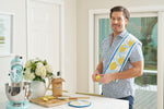 A man stands in a bright kitchen with the Lemon Kitchen Bundle—featuring a lemon-patterned kitchen towel, pot holder, and spatula—over his shoulder, next to a stand mixer, flowers, brown sugar, and sliced lemons.