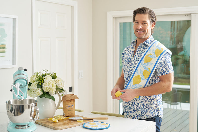 A man stands in a bright kitchen with the Lemon Kitchen Bundle—featuring a lemon-patterned kitchen towel, pot holder, and spatula—over his shoulder, next to a stand mixer, flowers, brown sugar, and sliced lemons.