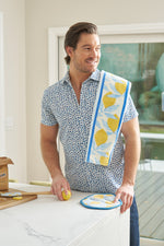 A man in a blue-patterned shirt stands smiling in a bright kitchen, holding a lemon near the Lemon Kitchen Bundle—featuring a lemon-print towel, potholder, and spatula—displayed on the counter beside packaged cookies.