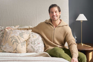 A man with brown hair and a beard, wearing a tan zip-up sweater and green pants, sits smiling on a bed featuring the Fall Florals Pillow. A small lamp and round wooden table are beside him.