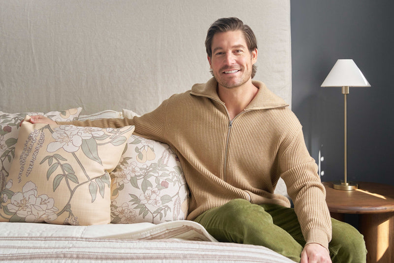 A man with brown hair and a beard, wearing a tan zip-up sweater and green pants, sits smiling on a bed featuring the Fall Florals Pillow. A small lamp and round wooden table are beside him.
