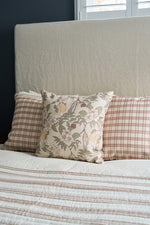 A neatly made bed features two plaid pillows and a Fall Florals Pillow in front, all in neutral tones. A beige headboard and sunlight streaming through white shutters complete the cozy, inviting scene.