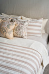 A neatly made bed with cream and brown striped bedding, two striped pillows, two floral-patterned pillows, and the Fall Pear Tree Pillow adds an autumn accent against a light headboard and wall.