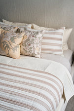 A neatly made bed with cream and brown striped bedding, two striped pillows, two floral-patterned pillows, and the Fall Pear Tree Pillow adds an autumn accent against a light headboard and wall.