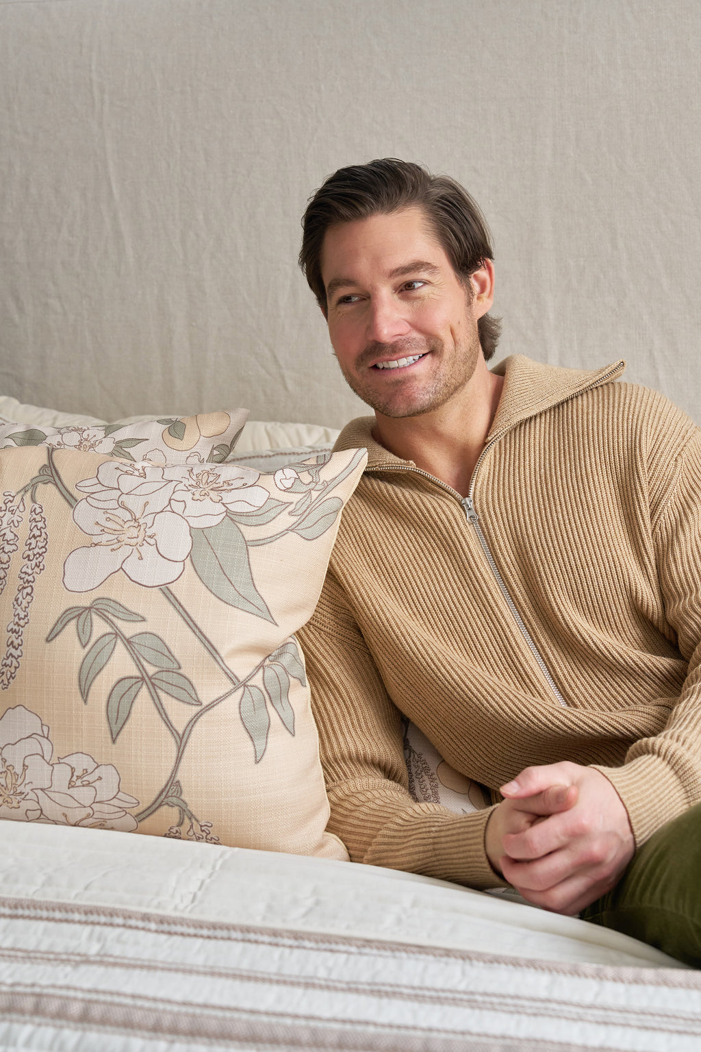 A man with short brown hair and a trimmed beard, wearing a tan zip-up sweater, sits on a bed with neutral bedding, smiling as he looks to the side beside the Fall Pear Tree Pillow, which adds an autumn accent to the room.