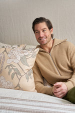 A man with short brown hair and a trimmed beard, wearing a tan zip-up sweater, sits on a bed with neutral bedding, smiling as he looks to the side beside the Fall Pear Tree Pillow, which adds an autumn accent to the room.