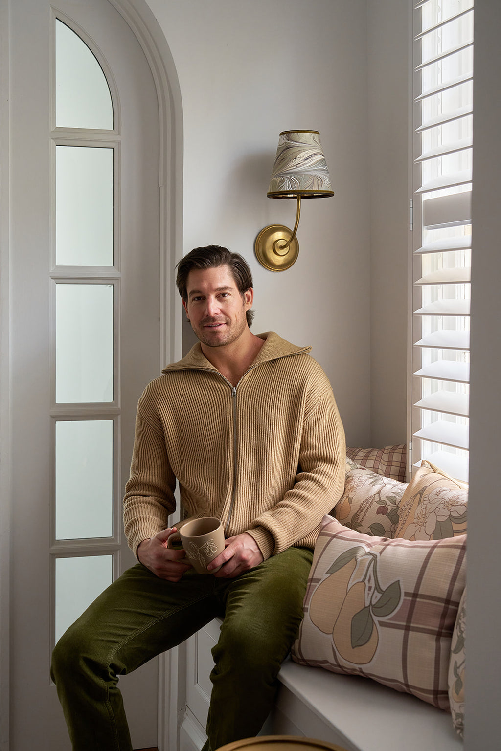 A man in a beige zip-up sweater and green pants sits on a window seat with a mug, surrounded by the Fall Pillow 4 Pack: Get 1 of Each! as sunlight streams in through white blinds.