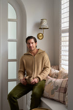 A man in a beige zip-up sweater and green pants sits on a window seat with a mug, surrounded by the Fall Pillow 4 Pack: Get 1 of Each! as sunlight streams in through white blinds.