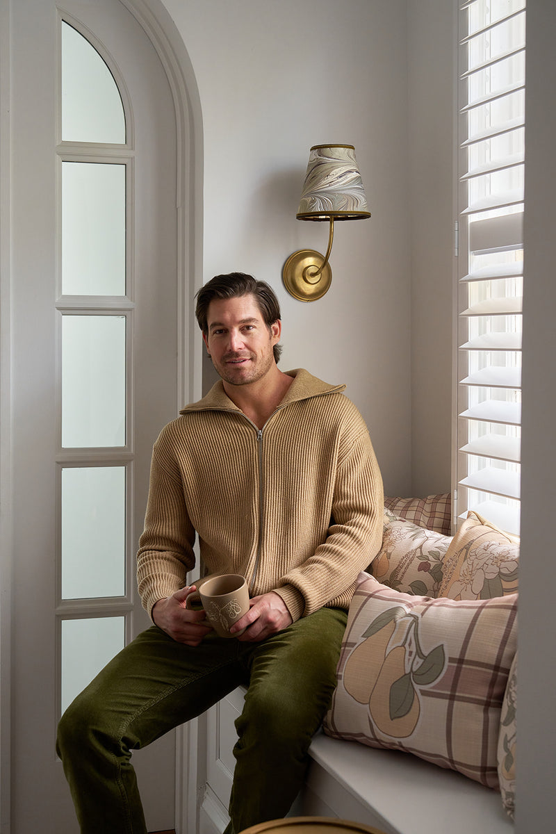 A man in a beige zip-up sweater and green pants sits on a window seat with a mug, surrounded by the Fall Pillow 4 Pack: Get 1 of Each! as sunlight streams in through white blinds.
