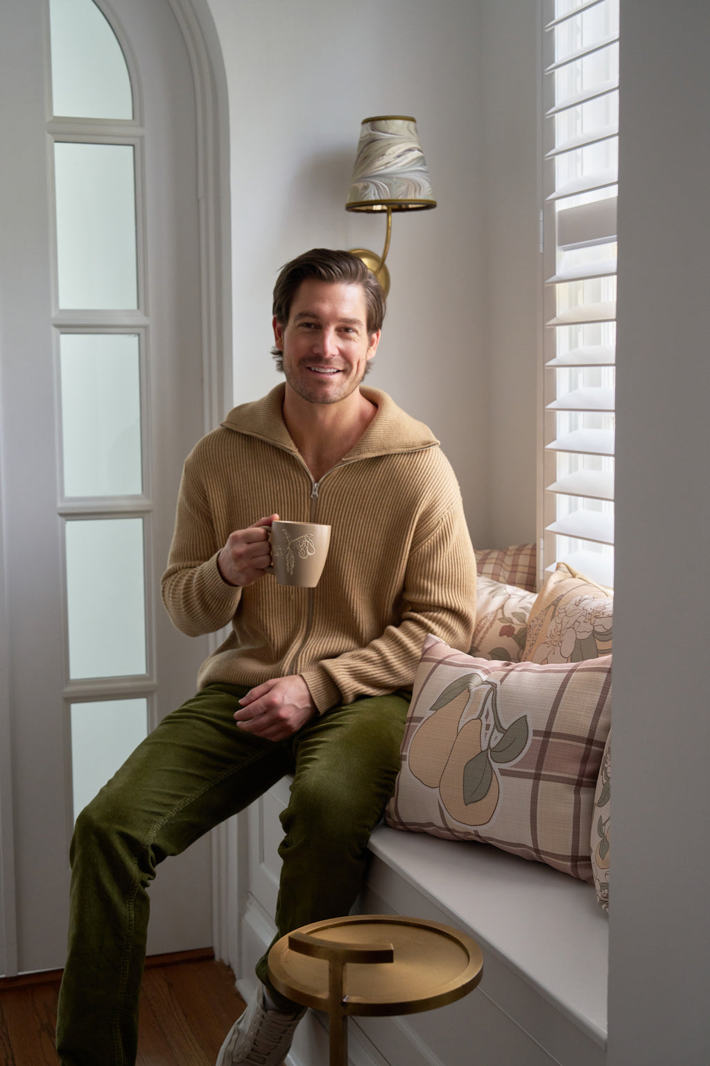 A man in a beige sweater and green pants smiles on a window seat, holding a mug. Sunlight streams through white shutters, highlighting cozy autumn decor with patterned pillows like the Fall Tartan Plaid Pillow.