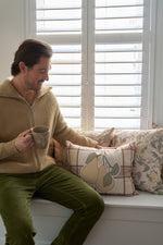 A brown-haired man in a tan zip-up sweater and green pants sits on a window seat with a mug, looking at decorative pillows, including the Fall Pears on Tartan Plaid Lumbar Pillow, in front of white shutters.