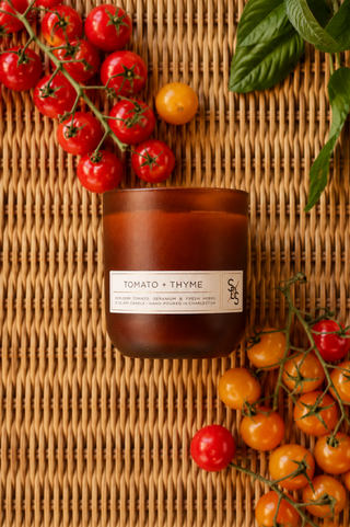 A Tomato + Thyme Limited Edition Candle in a brown jar sits on a woven mat, surrounded by red and yellow cherry tomatoes and fresh basil leaves.