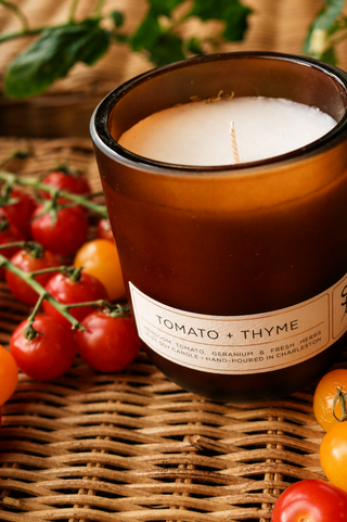 The Tomato + Thyme Limited Edition Candle, in brown glass, rests on a woven surface among red and yellow cherry tomatoes, with green vines and leaves behind it.