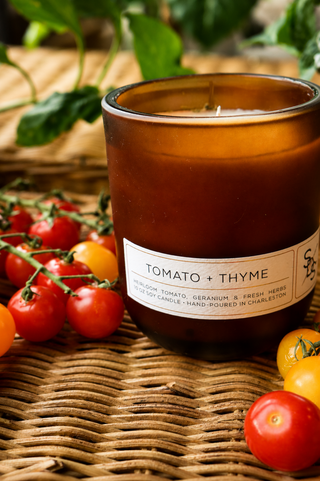 The Tomato + Thyme Limited Edition Candle in brown glass rests on a woven surface, surrounded by fresh cherry tomatoes and green leaves.
