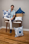 A man sits on a bench, surrounded by decorator-quality pillows in blue and white patterns. He holds the Modern Collection: Navy Coral Pillow from Sewing Down South™. Nearby, a basket teems with coastal chic pillows on the wooden floor against a neutral wall.