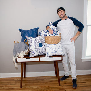 A man in a baseball cap and casual clothes stands by a wooden table topped with Navy Coral Oversized Pillows, a basket, and fringed blanket in a bright room with hardwood floors.