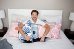 A man sits on a bed holding a Navy Coral Pillow from the Modern Collection by Sewing Down South™, surrounded by decorator-quality pillows in vibrant red. The coastal chic room features a neutral palette and sleek white bedside lamps.