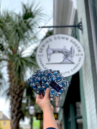 A hand displays blue floral fabric masks by a Sewing Down South shop sign, while a Crab Craze Koozie 4 PACK (Skinny or Standard) brings coastal flair. A palm tree and building are visible in the background.