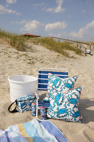 A coastal beach scene includes a white bucket, a blue and white striped chair, and two Crab Craze Shades of Blue Outdoor Pillows. A matching insulated bag sits next to two cylindrical containers in the sand, while a colorful towel lies against sand dunes beneath a cloudy sky.