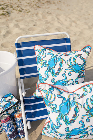 A beach scene with a blue-striped chair and two vibrant Crab Craze pillows adds coastal flair. A white cooler and canned drinks feature against a sandy, grassy backdrop. The Sewing Down South™ pillows add charm to the setting.