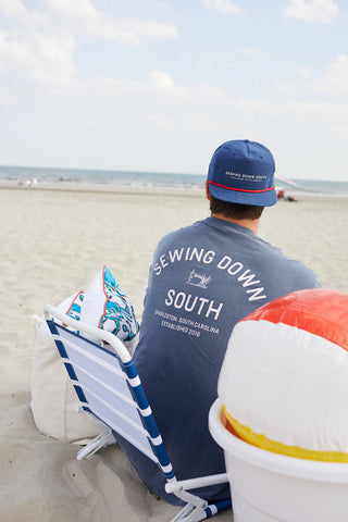 A person relaxes on a beach chair facing the ocean, wearing an SDS Vintage Logo Long-Sleeve Tee in Denim and a matching cap. A colorful beach ball and patterned pillow sit beside them on the sand.