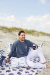 A man relaxes on a patterned blanket at the beach, holding a can in a koozie. Hes wearing an SDS Vintage Logo Long-Sleeve shirt in denim and white pants, surrounded by nautical-design cushions. The partly cloudy sky and grassy dunes create the perfect backdrop for relaxation.