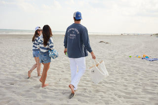Three people in casual beachwear, one wearing the SDS Vintage Logo Long-Sleeve Tee in Denim, walk barefoot on sandy beach with bags and towels. The ocean and cloudy sky form the backdrop, with scattered beach toys on the sand.