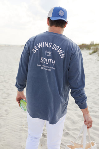 Strolling the beach with a drink and beige tote, a person sports the SDS Vintage Logo Long-Sleeve Tee in Denim and a blue cap. The ocean backdrop highlights this unisex garment-dyed cotton shirt from Sewing Down South.