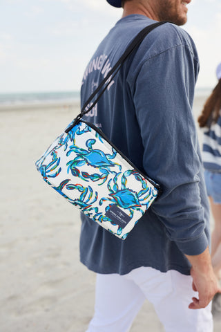 A person wearing the SDS Vintage Logo Long-Sleeve Tee in Denim and white pants stands on a beach, carrying a small cooler bag with a blue crab pattern. The ocean and another person appear in the background.