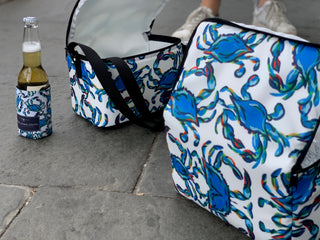 Two insulated cooler bags and a Crab Craze Koozie 4 PACK (Skinny or Standard), all featuring a blue crab pattern and durable neoprene, sit on gray stone pavement. One cooler is open, showing its white interior by some white sneakers.