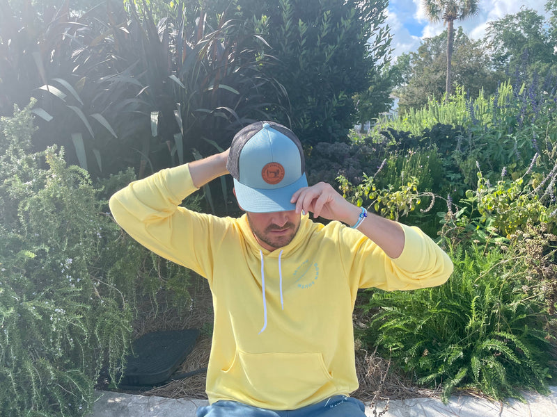 Wearing a blue Sewing Down South Leather Patch Trucker with black mesh, a person lounges outside on a sunny day amidst lush greenery and plants, under a clear blue sky.