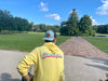 A person wearing a yellow Charleston hoodie and a Sewing Down South Leather Patch Trucker, Blue + Black Mesh cap is sitting on a bench, gazing at the open grassy park dotted with trees beneath a blue sky brushed with white clouds.