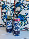 Two drinks, one bottled and one canned, are wrapped in Crab Craze Koozies from the 4 PACK (Skinny or Standard), featuring blue crab designs. They sit on a white surface in front of pillows with matching patterns.