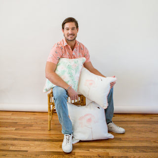 Wearing a pink patterned shirt and jeans, a person sits on a wicker chair holding The Flamingo Lumbar Pillow by Sewing Down South. The scene features a plain white wall and wooden floor, highlighting the charm of this Southern artist collaboration.