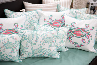 Discover the Outdoor Coral King Crab Pillow, showcasing vivid crab and seashell designs in pastel pink, teal, and green. Resting on a teal fabric surface, its paired with a striped pillow for a stylish addition to any space.
