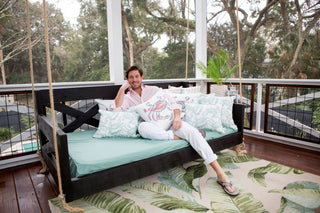 A person in a light pink shirt and white pants sits on a porch swing bed adorned with Pastel Crab Craze Outdoor Pillows. The porch, overlooking trees and a pool, features a rug with leaf patterns, creating an effortlessly inviting and serene ambiance.