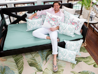 A person in a light pink shirt and white pants lounges on a swing bed with Pastel Crab Craze Outdoor Lumbar Pillow. Resting their arm on the pillow and wearing flip-flops, they enjoy the comfort of SDS Shield® Outdoor Fabric, while a leaf-patterned rug covers the floor.