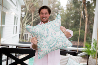 A man in a pink shirt smiles while holding the Pastel Crab Craze Outdoor Pillow, crafted with SDS Shield® Outdoor Fabric. He stands on a porch, with blurred trees behind him adding to the serene atmosphere of this charming Sewing Down South™ design.
