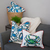 A gray sofa features two Crab Craze, Shades of Blues pillows, one rectangular and one square. A white knitted blanket enhances the coastal vibe. Beside it sits a basket with additional crab-themed pillows from Sewing Down South and a potted plant.