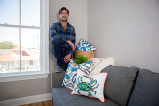 A man in a blue plaid shirt crouches near a window, grinning. Next to him, a gray sofa is adorned with vibrant crab-patterned pillows, featuring The King Crab Lumbar Pillow by Sewing Down South. A woven basket with a plant and a cream blanket complete the cozy décor.