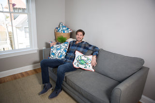 A man sits on a gray couch in a bright living room with light walls and a window. He wears a plaid shirt and jeans, surrounded by colorful Crab Craze, Shades of Blues Pillows from Sewing Down South™. Natural light enhances the coastal flair of the space.