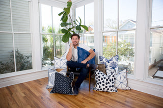 A man sits in a sunlit room on a chair, surrounded by coastal chic decorative pillows, including the Navy Medallion Oversized Pillow featuring the signature Sewing Down South™ pattern. Large windows and a tall green plant are in the background.
