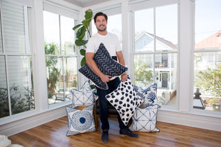 A man stands in a bright room with large windows, holding three decorative throw pillows featuring blue and white patterns, including the Navy Medallion Oversized Pillow. More coastal chic pillows are stacked at his feet beside a tall plant in the corner.