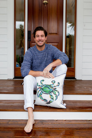A man sits barefoot on a wooden porch, leaning against a white door. Wearing a blue sweater and white pants, he smiles while holding an Outdoor King Crab Pillow with Navy, made from SDS Shield® Outdoor Fabric with a painted crab design.