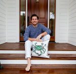A man sits barefoot on a wooden porch, leaning against a white door. Wearing a blue sweater and white pants, he smiles while holding an Outdoor King Crab Pillow with Navy, made from SDS Shield® Outdoor Fabric with a painted crab design.