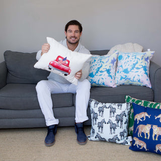 A person sitting on a gray couch holds up a throw pillow with a red truck design. The couch is adorned with the Giraffes on Grass Lumbar Pillow and others featuring animal patterns like zebras and tigers, adding an exotic flair. The person is dressed in a white sweater and pants.