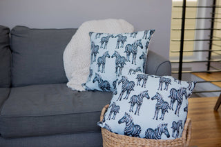 A gray couch displays exotic flair with a white knitted throw over the back. Two Giraffes on Grass Lumbar Pillows add bold animal patterns, while another pillow in a woven basket rests on the floor. The room features a soft, neutral color scheme.