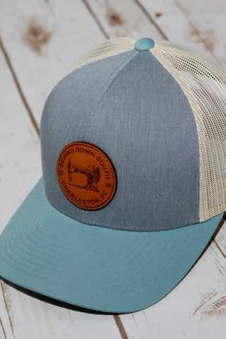 A Sewing Down South Leather Patch Trucker cap in grey with a blue front, light blue bill, and white mesh back rests on a light wooden surface. The front displays a brown leather patch with vintage logo text next to a sewing machine graphic.
