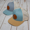 On a wooden surface rest two Sewing Down South Leather Patch Truckers. One is light blue (Smoke Blue), and the other has a gray and cream mix, both with tan brims, mesh backs, circular leather patch logos on the front panel, and adjustable snapbacks for an ideal fit.