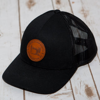 The Sewing Down South Leather Patch Trucker in black on black features a leather patch with embossed text and a South Carolina outline, combining style and comfort in this mesh snapback. It sits on a light wooden surface.